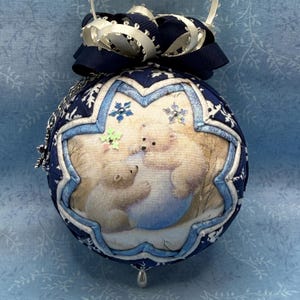May include: A round Christmas ornament with a navy blue and white snowflake pattern. The ornament features a central image of two polar bears. A navy blue bow and silver chain complete the festive design.