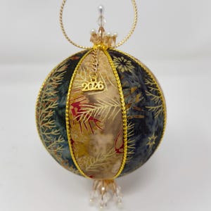 May include: A handcrafted Christmas ornament with a gold hanging loop and beaded accents. The spherical ornament features panels of fabric in shades of blue, gold, and red, with a gold trim. A small charm with the year "2026" hangs from the top.