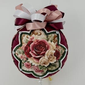 May include: A spherical fabric ornament featuring a central panel with a floral rose print in shades of red, cream, and green. The ornament is bordered by dark burgundy fabric folds, accented with small white beads, and topped with a multi-layered bow.