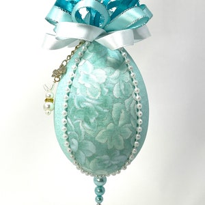 May include: A light blue fabric Christmas ornament with a white floral pattern, trimmed with white pearls and a blue and white ribbon.