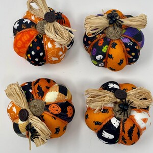 May include: Four small fabric pumpkins with Halloween patterns. Each pumpkin features orange, black, and white fabrics, adorned with raffia, a button, and a small spider. These pumpkins are designed for seasonal decoration.