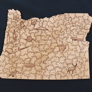 May include: A wooden jigsaw puzzle of the state of Oregon, with the names of cities and towns throughout the state. The puzzle is made of light brown wood and has a natural finish. The puzzle is in the shape of the state of Oregon and includes various icons representing the state, such as a beer mug, a fish, a mountain, and a deer.