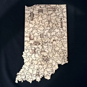 May include: A wooden jigsaw puzzle shaped like the state of Indiana, with various cities and landmarks depicted throughout the puzzle. The puzzle is made of light brown wood and features a variety of illustrations, including the state capitol building, a racing car, and a cornfield.