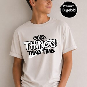 Könnte beinhalten: Ein beiges T-Shirt mit kurzen Ärmeln und einem schwarz-weißen Grafikdruck auf der Brust mit dem Text GOOD THINGS TAKE TIME in einer fetten, stilisierten Schrift. Ein kreisförmiges schwarzes Abzeichen oben rechts zeigt den Text Premium Bügelbild.