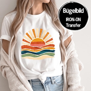Könnte beinhalten: Weißes T-Shirt mit einem Retro-Sonnenuntergangsdesign in Orange, Gelb, Rot und Blau. Die Grafik zeigt eine Sonne mit Strahlen und geschichteten Wellen. Der Text "IRON-ON TRANSFER" ist in einem schwarzen Kreis zu sehen.