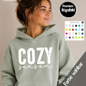 Könnte beinhalten: Ein salbeigrüner Kapuzenpullover mit dem weißen Schriftzug COZY season auf der Vorderseite. Das Design enthält eine kleine kreisförmige Grafik in der oberen rechten Ecke sowie eine Farbkarte mit verschiedenen verfügbaren Farboptionen.