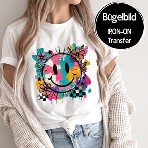 Könnte beinhalten: Weißes T-Shirt mit einem farbenfrohen Smiley-Gesicht-Design, Blumen und Sternen. Das Design beinhaltet ein Schachbrettmuster und den Text "IRON-ON TRANSFER" in einem schwarzen Kreis. Das Shirt ist ein lässiger Stil.