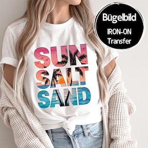 Könnte beinhalten: Weißes T-Shirt mit den Worten "SUN SALT SAND" in farbenfrohem, stilisiertem Text. Das Design zeigt eine Sonnenuntergangsszene mit Palmen und Vögeln. Ein schwarzer Kreis mit der Aufschrift "Bügelbild IRON-ON Transfer" befindet sich oben rechts.