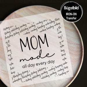 Könnte beinhalten: Eine transparente Bügelbild-Transferfolie mit dem schwarzen Text MOM mode all day every day in der Mitte, umgeben von einem sich wiederholenden Muster der Wochentage in Schreibschrift, auf einer hellen, runden Holzoberfläche präsentiert.