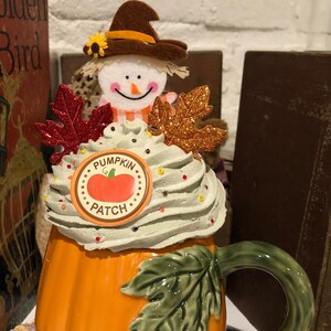 May include: Orange pumpkin-shaped ceramic mug with a green leaf and a white whipped cream top. The mug is decorated with a scarecrow wearing a brown hat, red and gold glitter leaves, and a round sticker that says "Pumpkin Patch" with a small pumpkin image.