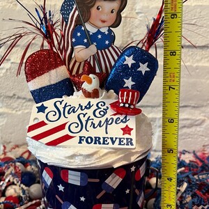 May include: A patriotic centerpiece with a blue and white popsicle-themed bucket, topped with a doll holding a flag, glittery stars, and a sign that reads "Stars & Stripes Forever." The height is approximately 9 inches.