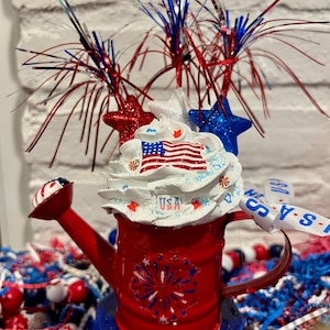 May include: A red watering can decorated with patriotic elements. It features a white whipped cream topping with an American flag design, red and blue stars, and firework-like accents. The base is blue, and the handle has a white ribbon with the text "USA".