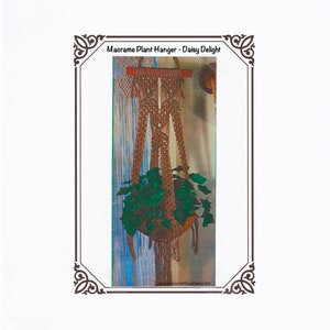 May include: Macrame plant hanger, "Daisy Delight", with a woven design in shades of brown. The plant holder suspends a basket filled with green leafy plants. The text "Macrame Plant Hanger - Daisy Delight" is at the top.
