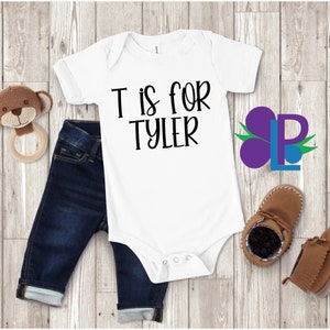 May include: White baby onesie with black text that reads "T is for Tyler". The onesie is laying on a wooden surface with a pair of blue jeans, a brown teddy bear, and a pair of brown baby shoes.