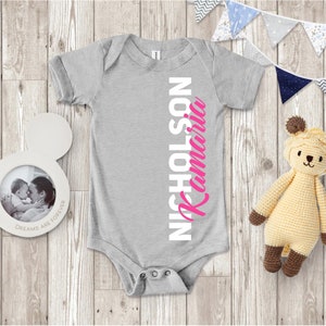 May include: A gray baby onesie with the name "Kamari Nicholson" printed vertically down the right side. The name "Kamari" is in pink and the name "Nicholson" is in white.