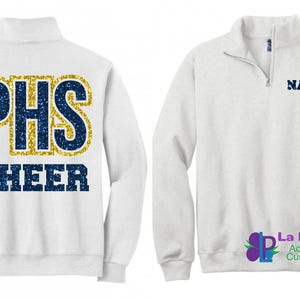 May include: White quarter-zip sweatshirt with "PHS CHEER" in navy blue and gold glitter lettering on the back. The front features a navy blue "NAME" text near the collar. A logo is visible on the lower right.