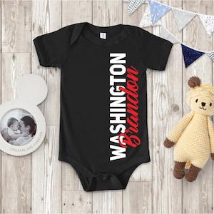 May include: Black baby onesie with the name "Washington Brandon" printed vertically in white and red. The onesie is short-sleeved and has a snap closure. A photo frame and a stuffed bear are next to the onesie.