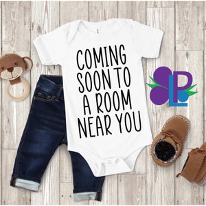 May include: White baby onesie with black text that reads "Coming soon to a room near you". The onesie is laid out on a wooden surface with a pair of blue jeans, a brown teddy bear, and brown baby shoes.