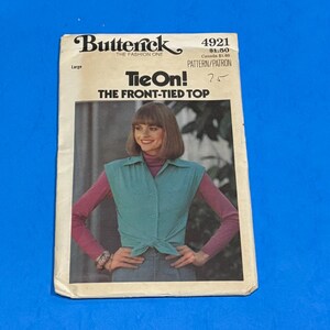 May include: A vintage Butterick sewing pattern for a front-tied top. The pattern cover features a woman wearing a teal top tied at the waist over a pink long-sleeved shirt and blue jeans. The text on the cover reads "Tie On!" and "The Front-Tied Top."