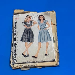 Peut inclure: Un ancien patron Simplicity à 15¢, taille 10, avec des illustrations de deux robes. L'une est à carreaux noirs et blancs, l'autre bleu clair. Le patron est sur fond bleu.