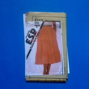 May include: A stack of vintage sewing patterns on a blue background. The top pattern features a photo of an orange skirt. The text on the pattern reads "Simplicity 5113" and "Extra-Sure Pattern".