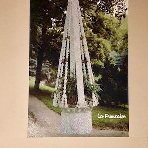 May include: A white macrame plant hanger with a glass bowl holding greenery. The hanger has a conical shape with wooden beads and a fringed bottom. The image has the text "La Francaise".