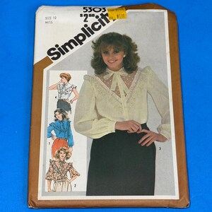 May include: A Simplicity 5303 sewing pattern envelope for a size 12 miss blouse. The cover features an illustration of a long-sleeved cream blouse with a sheer lace yoke, a tie-neck collar, and ruffles, paired with a solid black skirt.