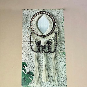 May include: A circular macrame mirror with a cream-coloured woven design and a small, oval mirror at the centre. The macrame features brown and cream-coloured accents and hanging tassels. The mirror is displayed on a textured, grey and white background.