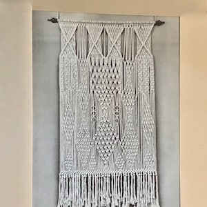 Puede incluir: Tapiz de pared de macramé blanco con detalles geométricos de diamantes y flecos. La pieza de arte textil hecha a mano presenta una serie de formas de diamantes y un borde inferior con flecos. La decoración de la pared está montada en una barra de metal oscuro.
