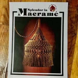 May include: A printed page with the title "Splendor in Macramé" featuring a macramé lamp design. The lamp is a warm brown color with a fringed bottom and intricate knotting. The background is a gradient of dark red to black.