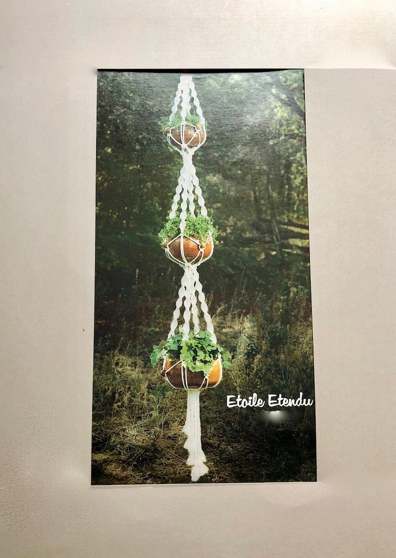 May include: A macrame plant hanger with three wooden bowls filled with green plants. The white macrame cord is knotted to create a decorative design. The background shows a blurred forest scene. The text "Etoile Etendu" is visible.