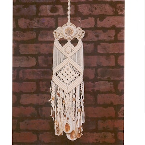 May include: Cream-coloured macrame wall hanging with a decorative top featuring a sun-like design. The intricate knotted pattern includes diamond shapes and beaded accents, with hanging tassels and shell-like pendants. A bohemian-style home decor item.