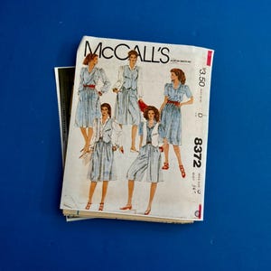 May include: A vintage McCall's sewing pattern featuring illustrations of women's clothing designs. The pattern cover displays various outfits, including dresses and jumpsuits, in a light blue fabric with red belts. The pattern number is 8372.