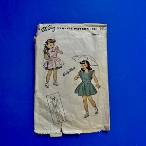 May include: Vintage De Barry Perfect Pattern size 6 sewing pattern. The pattern features illustrations of a young girl wearing dresses. The pattern is printed on aged paper with the text "Easy-Made". The background is a solid blue.