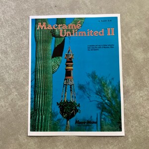 May include: A book cover titled "Macrame Unlimited II" featuring a macrame plant hanger with a green plant inside. The background shows a desert scene with a cactus and blue sky. The book is by Macra-Weave.