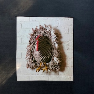 May include: A handmade turkey wall hanging. The turkey is made of brown and tan yarn, with a red wattle and yellow feet. The background is a white brick pattern.