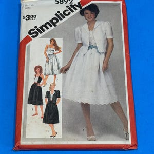 Puede incluir: Un sobre de patrón de costura Simplicity 5892 para talla 12. La portada presenta ilustraciones de un vestido blanco con chaqueta a juego y fajín azul, un vestido negro con corpiño blanco y un vestido negro con chaqueta negra sobre corpiño.