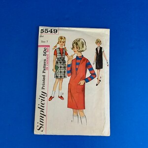 May include: Vintage Simplicity dressmaking pattern for a girl, size 7. The cover displays illustrations of three dress designs. The central image shows a girl in a red dress with pockets. The cover text includes "Simplicity" and "Printed Pattern".