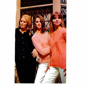 May include: Three women in vintage clothing pose outdoors. One wears a black button-down dress with a floral brooch. The other two are in peach-colored knit sweaters and white pants. The image has a retro, fashion-forward aesthetic.
