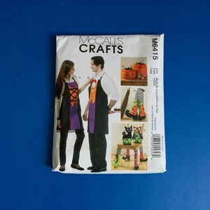May include: A McCall's Crafts pattern envelope against a blue backdrop. The envelope shows Halloween-themed aprons and decorations, including pumpkins, witch legs, and owl figures. The pattern number is M6415.
