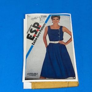 Può includere: Un cartamodello Simplicity E.S.P. Extra-Sure Pattern 5581 per le taglie 10-12-14. La busta mostra un abito blu senza maniche con scollo quadrato, una cintura bianca e chiusura laterale con bottoni. Il testo riporta Adjustable for Miss Petite.