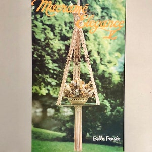 May include: A macrame plant hanger with a decorative basket holding dried flowers and crystals. The beige macrame design features wooden beads and a glass shelf. The image includes the text "Macrame Elegance II" and "Belle Pensée".