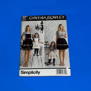 Puede incluir: Un patrón de costura Simplicity con diseños de Cynthia Rowley. La portada del patrón muestra a mujeres y niños con delantales y faldas. La portada es blanca con texto negro y un código de barras.