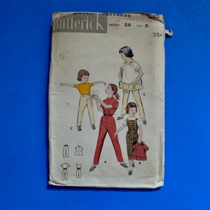 May include: Vintage Butterick sewing pattern featuring illustrations of children's clothing designs. The pattern includes images of a jumpsuit, overalls, and a dress. The pattern is for a breast size of 26 and a size 8. The pattern is on a blue background.