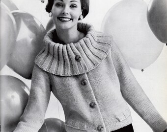 May include: A light-colored, knitted cardigan with a wide, ribbed collar and three large buttons. The cardigan has long sleeves and small pockets. The woman is wearing the cardigan with a black skirt, and is smiling in front of a background of balloons.