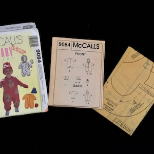 May include: Three McCall's baby wear sewing patterns. The main pattern shows a baby in a red jumpsuit with bear ears. The other patterns show diagrams and instructions for baby clothing. The patterns are on a black background.