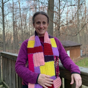 May include: A colorful, patchwork crochet scarf in shades of pink, purple, yellow, and red. The scarf is worn around the neck and chest. The person is wearing a purple fleece jacket.