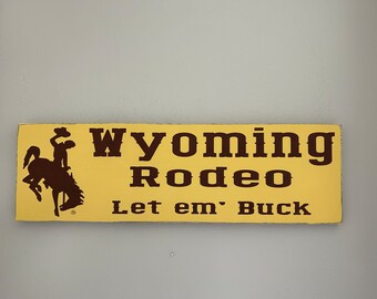 May include: A yellow sign with brown text and a silhouette of a cowboy riding a bucking horse. The sign reads "Wyoming Rodeo Let em' Buck".
