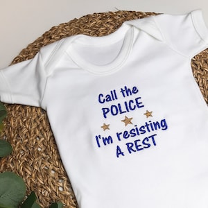 Può includere: Body bianco per neonato con testo blu che recita "Call the POLICE I'm resisting A REST" con tre stelle dorate sopra il testo.