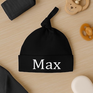 May include: A black baby beanie with the name "Max" in white block letters. The hat is knotted at the top and has a folded brim. Other baby items are in the background.